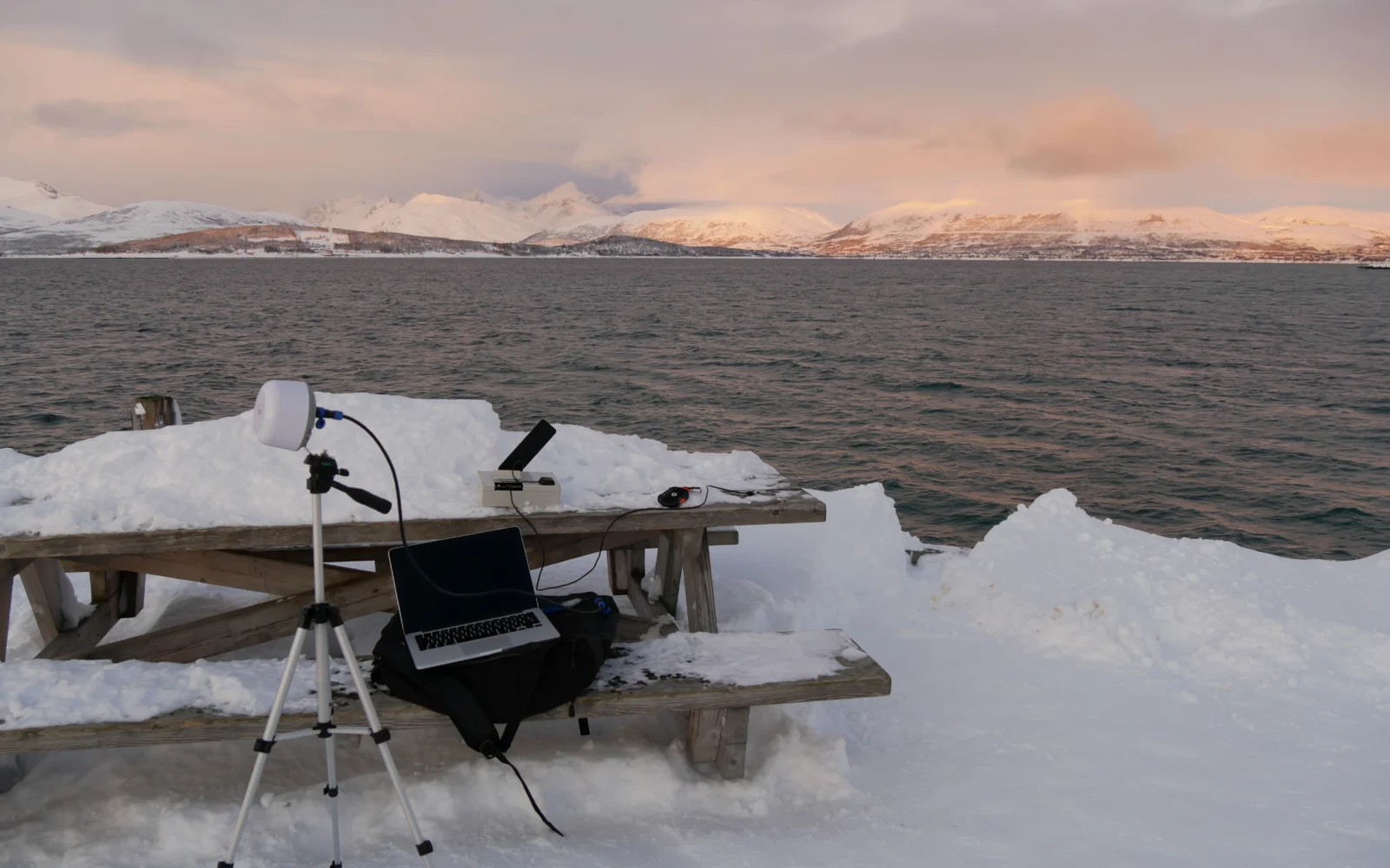 A laptop and white satellite antenna are set up on a snowy picnic table beside a calm body of water with pink-hued mountains in the background.