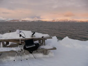A laptop and white satellite antenna are set up on a snowy picnic table beside a calm body of water with pink-hued mountains in the background.